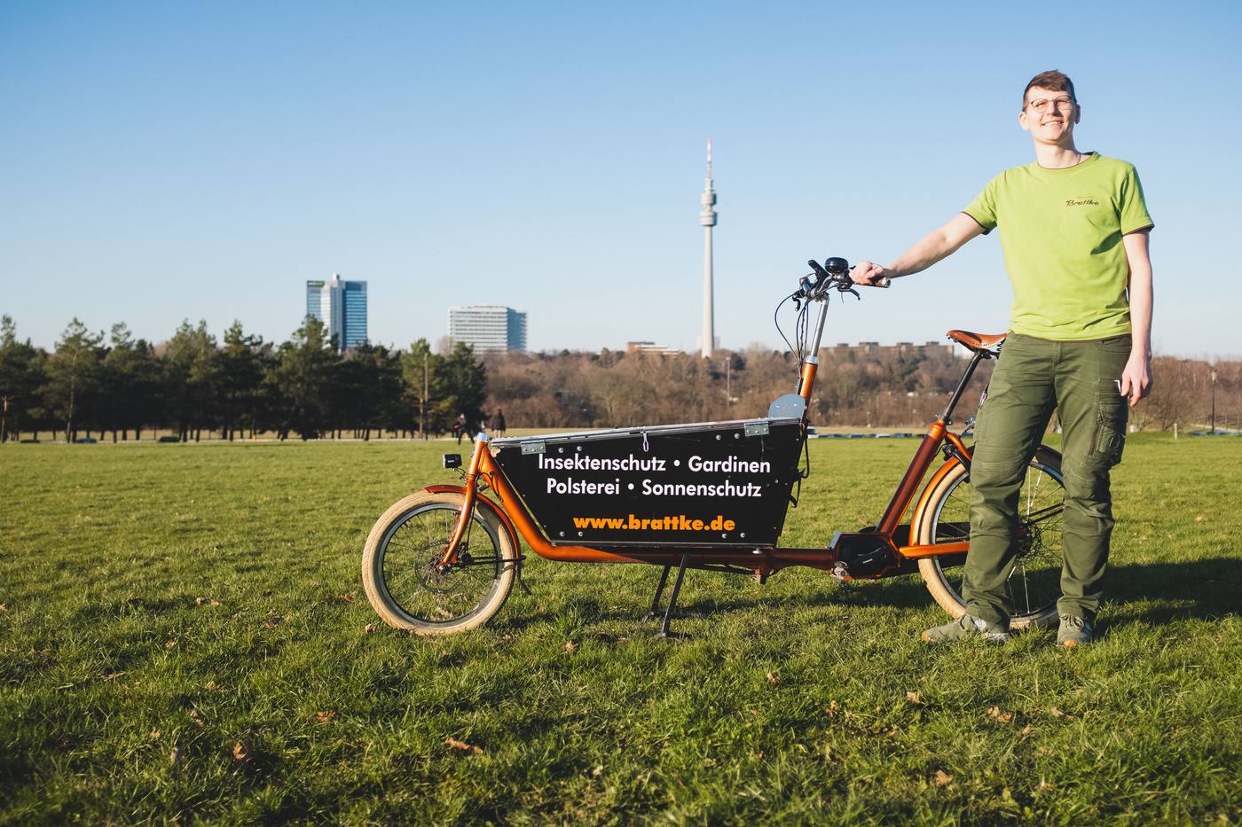 Knut Brattke steht neben dem orangen Lastenrad seines Betriebs, im Hintergrund der Florianturm in Dortmund